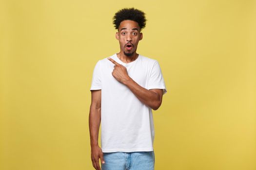 Astonished young African American man dressed in casual white shirt having excited fascinated look, pointing index finger at copy space on golden yellow background for your text or promotional content.