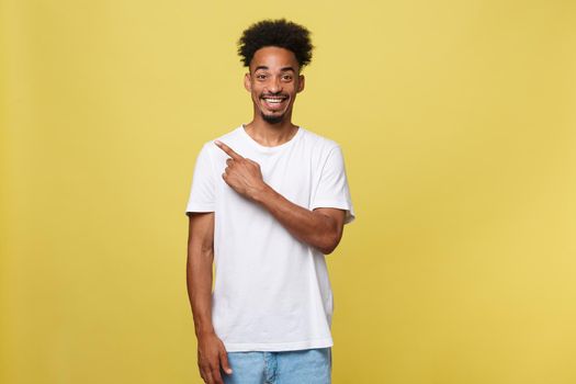 Astonished young African American man dressed in casual white shirt having excited fascinated look, pointing index finger at copy space on golden yellow background for your text or promotional content.