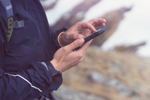 Man holds a smartphone in his hands and looks at a map, makes a route through the mountains, writes a message, chats. Young traveler explores the area, uses the app on his device while hiking.