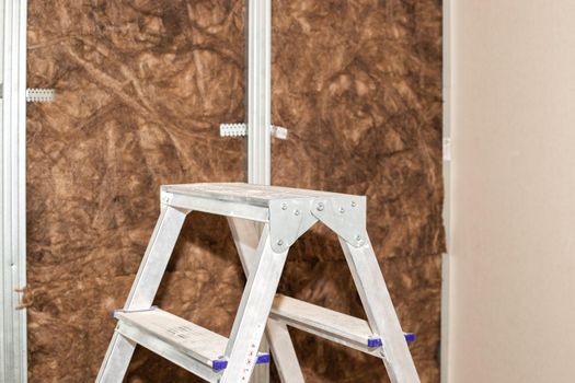 Home repairs. A folding ladder stands against the background of a wall lined with mineral wool for further cladding with plasterboard. Heat insulation and sound insulation of housing.