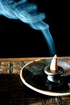 Image of A horned-looking piece of incense in a black tray burns blue smoke against a black background