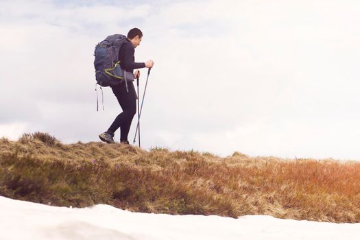 A traveler walks along a trail in the mountains, hiking in the wild in winter alone, with a backpack, camping equipment and trekking poles. The young man enjoys an active lifestyle.