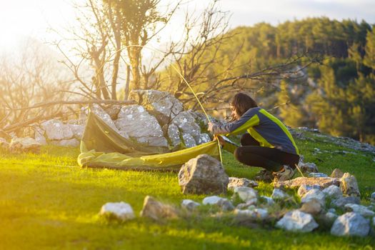 A young girl sets up her tent at a campsite on a green meadow among the mountains on a warm summer evening. The traveler is getting ready for a comfortable sleep in nature.