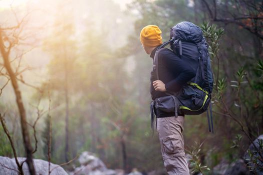 A young man with a backpack and camping equipment stands on the rocks and looks at the beautiful landscape in the sun, at the trees in a mountain gorge. Trekking the trail in the mountains.