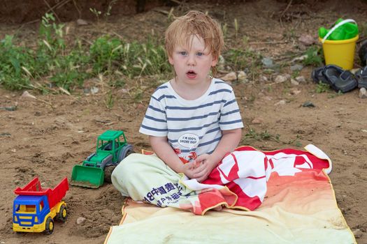 A little boy sits on a towel on a sandy beach among toys. Childrens holiday at the sea