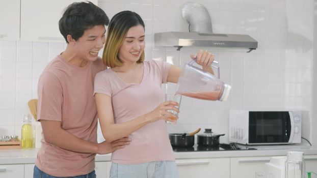 Happy Asian young family couple husband and wife enjoying drinking fresh apple smoothie juice in kitchen at home. woman poured juice from blender for man to drink. Healthy lifestyle.