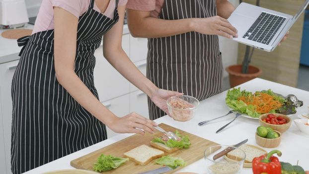 Happy Asian beautiful young family couple husband and wife cooking food vegetable salad in kitchen together at home. The man and woman according online cooking class to recipe on laptop computer