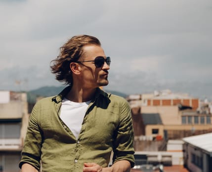 Handsome european man in sunglasses with long hair, standing outdoors and looking to the side
