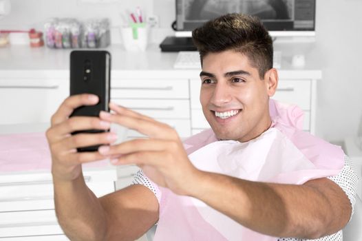 Happy patient taking selfie at dental clinic. Patient is holding smartphone and showing his healthy smile. High quality photo.