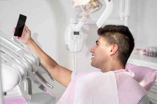 Happy patient taking selfie at dental clinic. Patient is holding smartphone and showing his healthy smile. High quality photo.