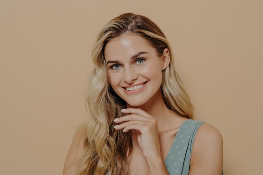 Close up portrait of lovely and cute blonde woman with blue eyes touching her chin with hand and looking at camera with broad smile and happy face expression while posing over beige background