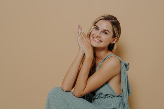 Pleasant young woman with natural makeup in elegant evening dress with open shoulders posing, happy female sitting on chair in studio background, smiling tenderly on camera with hands touching face