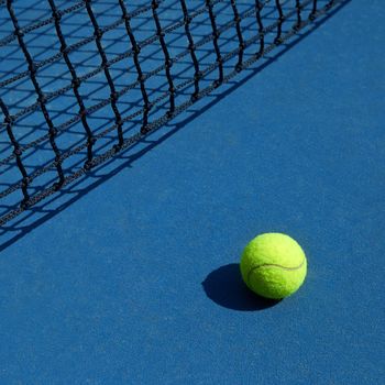 Yellow tennis ball is laying near black opened tennis court's net. Contrast image of sport equipment with satureted colors and shadows. Concept of tennis outfit photografing.