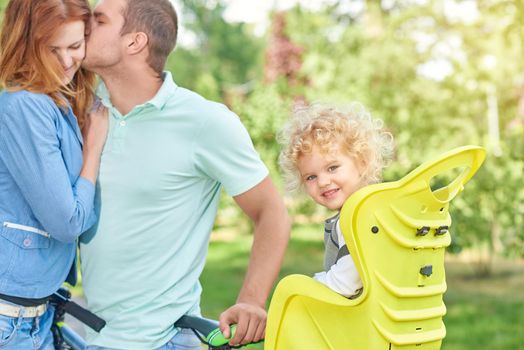 Adorable little girl smiling to the camera sitting in a baby bike seat while her parents cuddling on the background copyspace family love marriage happiness emotions childhood safety.