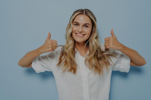Blonde female dressed in white shirt expressing positive emotions while showing thumbs up gesture with both of her hands, displaying that she likes it, isolated next to blue wall