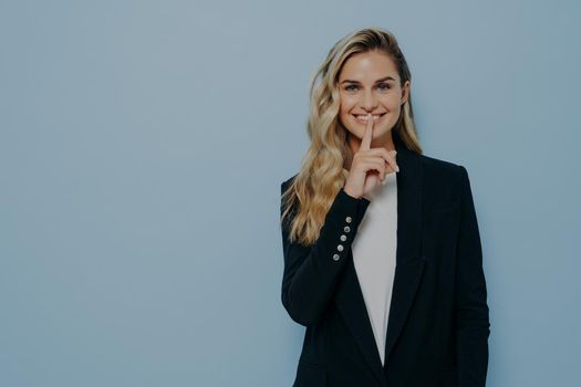 Cheerful beautiful young woman in black blazer keeps finger on lips and asking to keep something in secret or be silent while posing isolated against blue studio background, copy space for text