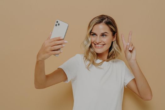 Cool joyful woman with blonde hair with smartphone in hand making selfie in studio against nude background and showing peace sign, taking picture of herself and smiling, dressed in white t shirt