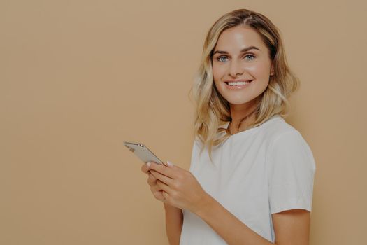Portrait of cheerful young woman in white tshirt being happy to read good news on smartphone, using cell phone and looking at camera with positive face expression. Modern technologies concept