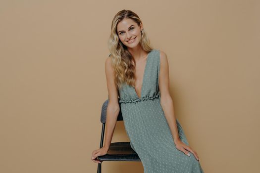 Young sensual and seductive blonde woman in long romantic summer dress posing over beige background in studio, sitting on black chair and looking at camera with love and satisfaction