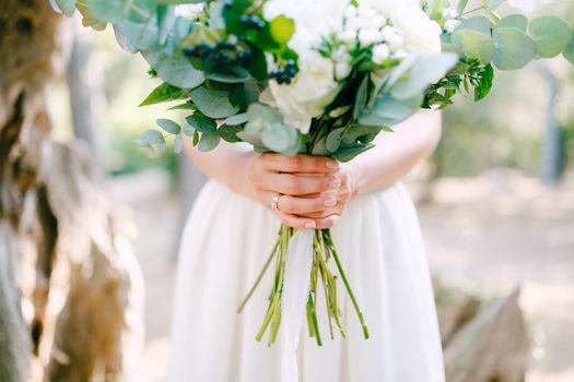The bride is holding a wedding bouquet of roses, eucalyptus branches, delicate white flowers and dark berries in her hands, close-up . High quality photo