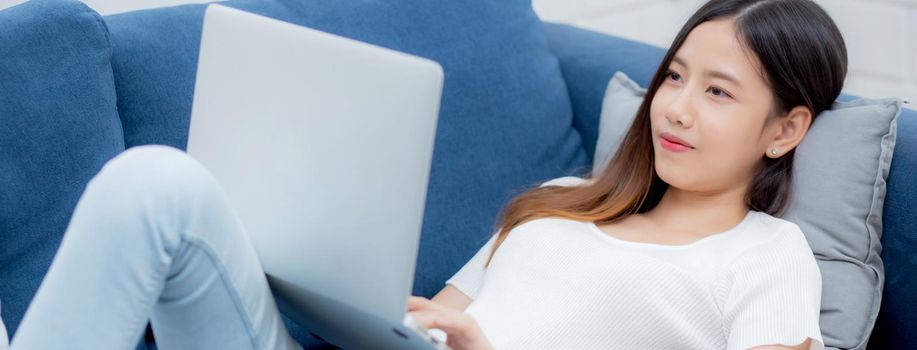 Young asian business woman smile and work from home with laptop computer online to internet on sofa in living room, freelance girl using notebook on couch with comfort, new normal, lifestyle concept.