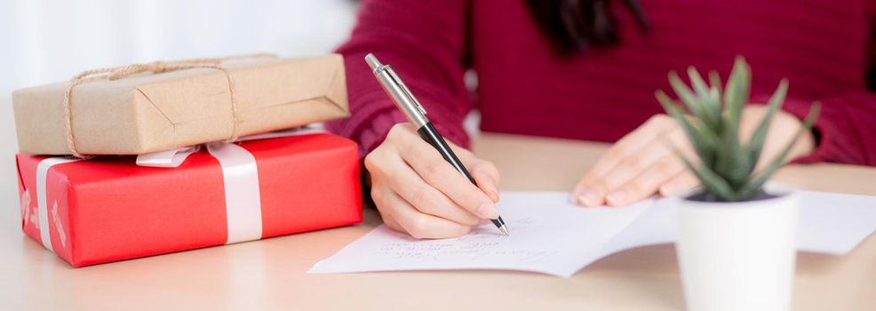 Closeup hand of young asian woman writing postcard in Christmas day at home, eve and celebrate, female writing message on greeting card with giving gift box in holiday, congratulation and celebration.