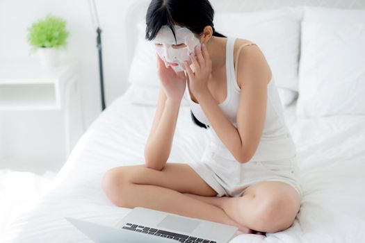 Beautiful young asian woman with sheet facial mask sitting working on laptop computer on bed at bedroom, beauty girl applying face mask for skincare of wrinkle at home, skin care, health and wellness.