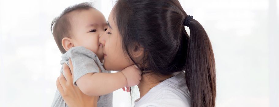 Young asian mother holding little baby girl and kiss together in the bedroom at home, mom carry child and care, woman and kid with carefree, toddler and parent, emotion and expression, family concept.