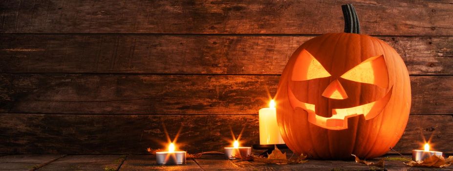 Halloween pumpkin glowing jack o lantern with carved face and candles on wooden background, banner with copy space for text