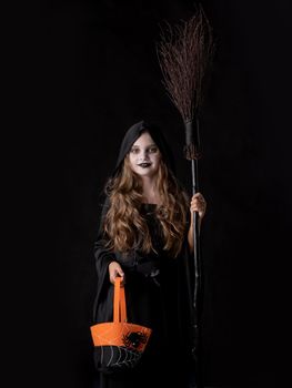 Girl in halloween witch costume hold broom and orange bag trick or treating isolated on black background