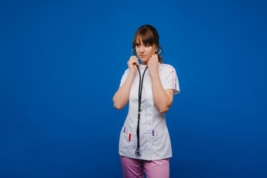 A female doctor, gesticulating, checks the heartbeat in the doctor's office at the hospital with a stethoscope isolated on a blue background.