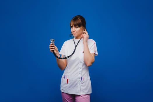 A female doctor, gesticulating, checks the heartbeat in the doctor's office at the hospital with a stethoscope isolated on a blue background.