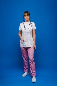 A female doctor, gesticulating, checks the heartbeat in the doctor's office at the hospital with a stethoscope isolated on a blue background.