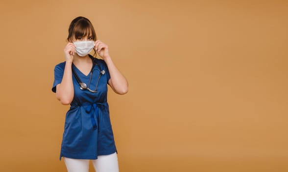 A girl doctor stands in a medical mask, isolated on a brown background.