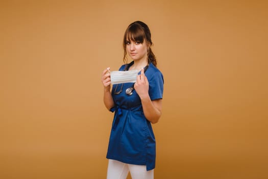 A girl doctor stands in a medical mask, isolated on a brown background.