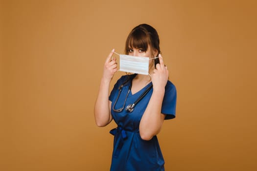 A girl doctor stands in a medical mask, isolated on a brown background.