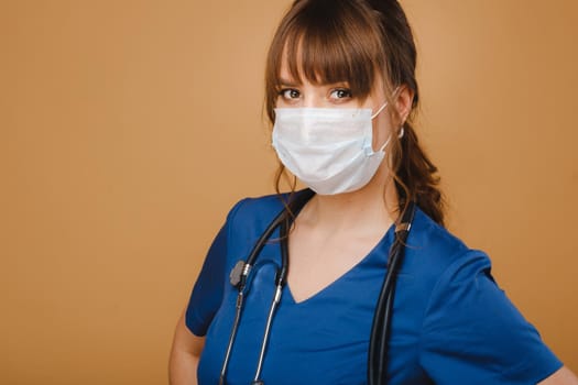 A girl doctor stands in a medical mask, isolated on a brown background.