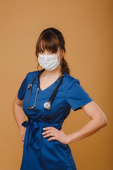 A girl doctor stands in a medical mask, isolated on a brown background.