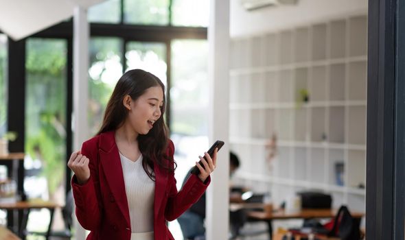 Smiling Asian woman holding mobile phone with fist hand and excited for success in office.