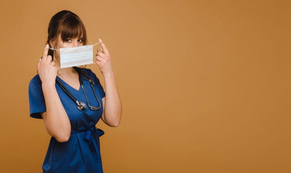 A girl doctor stands in a medical mask, isolated on a brown background.