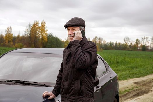 Adult pensioner stands near car and talks on smartphone against background of an autumn rural landscape. Rural life. Selective focus.