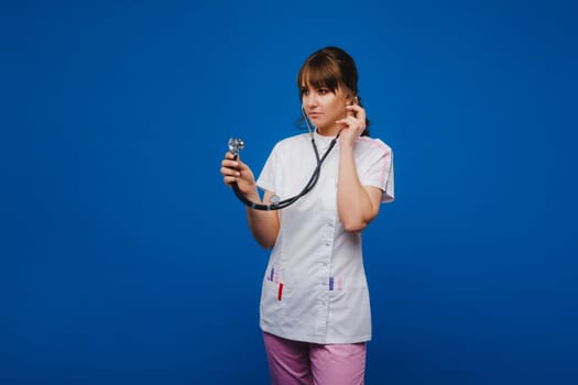 A female doctor, gesticulating, checks the heartbeat in the doctor's office at the hospital with a stethoscope isolated on a blue background.