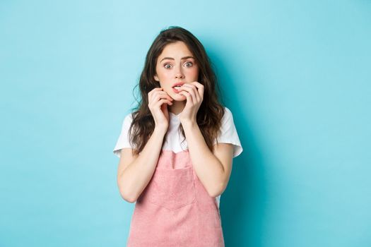 Portrait of scared and startled brunette girl biting fingernails, staring at camera shocked and worried, hear terrible news, standing in stupor against blue background.