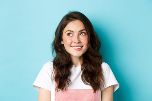 Close up of dreamy silly girl thinking of something, looking at upper left corner and smiling happy, daydreaming, staring at logo, standing over blue background.