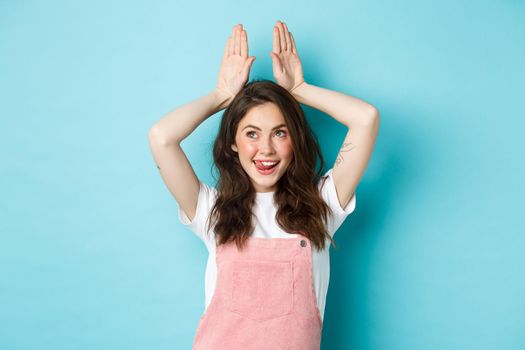 Image of happy young woman celebrating Easter, showing bunny ears with hands on head and smiling cheerful, enjoying spring holiday, standing over blue background.