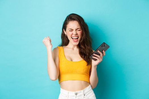 Technology and lifestyle concept. Satisfied woman achieve success, winning on mobile phone, making fist pump and scream yes with joyful expression, standing over blue background.