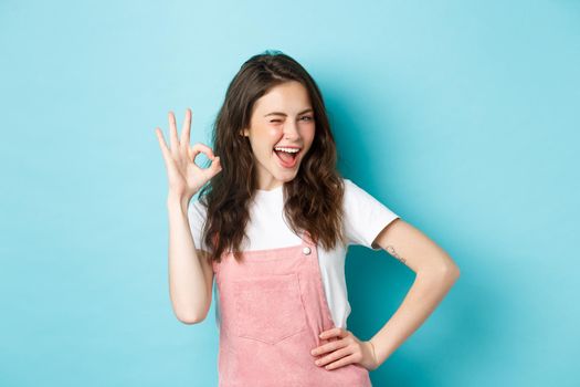 Image of cheerful glamour woman winking and showing OK sign to recommend and approve nice thing, like and agree, saying yes, praising good choice, blue background.