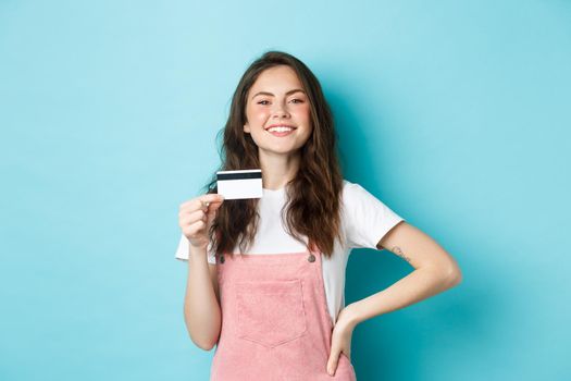 Proud smiling girl looking boastful, showing her plastic credit card, going to buy something, shopping contactless, standing over blue background.