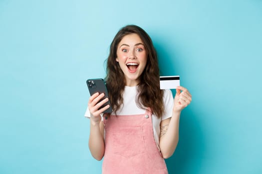 Surprised and happy young woman showing plastic credit card and smartphone, looking amazed at camera, shopping online with spring discounts, blue background.