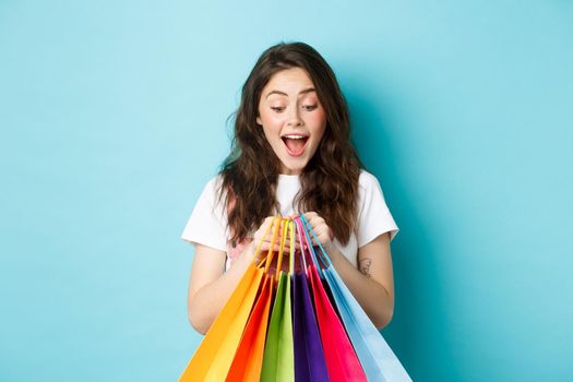 Happy young woman looking at her shopping bags with excitement, buying with discounts in shops, standing over blue background.